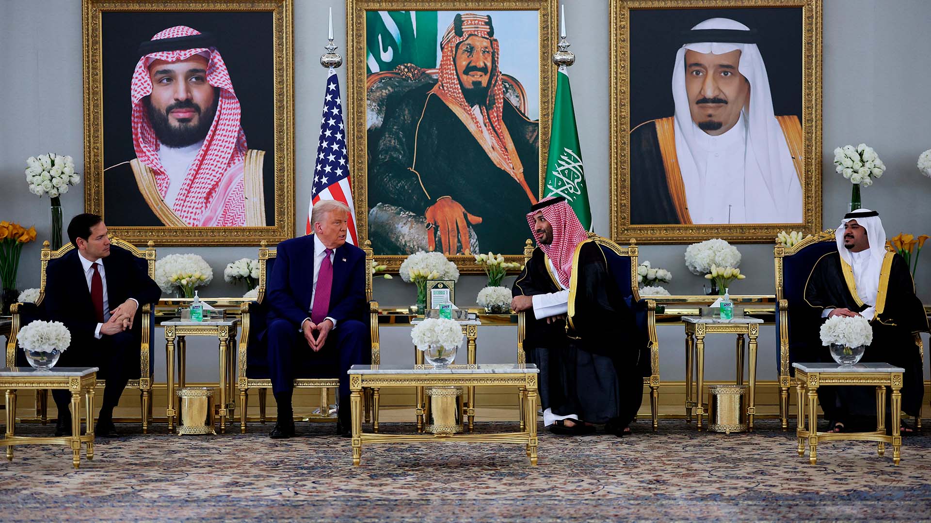 Donald Trump y el príncipe heredero saudí Mohammed bin Salman conversan sentados en una sala lujosamente decorada en Riad, con retratos de líderes saudíes al fondo, banderas de EEUU y Arabia Saudí tras ellos, y mesas doradas con arreglos florales. Trump