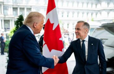 Donald Trump (de pie, con traje azul marino, corbata roja y camisa blanca) saluda con un apretón de manos a Mark Carney (también de pie, con traje y corbata azul marino y camisa blanca) en la entrada del Ala Oeste de la Casa Blanca, el 6 de mayo de 2025. En el centro de la imagen se aprecia una bandera de Canadá entre ambos. Al fondo se distingue parte del edificio y, a la derecha, un coche negro. Canadá