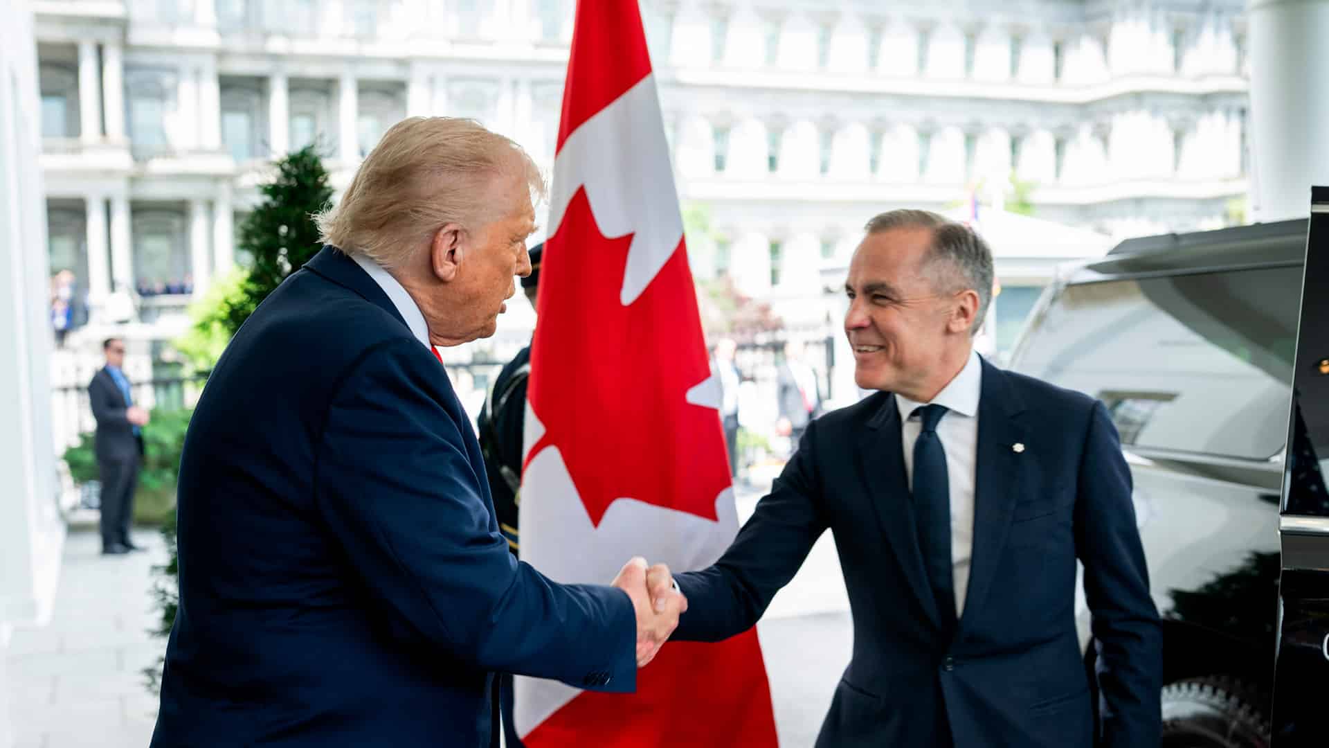 Donald Trump (de pie, con traje azul marino, corbata roja y camisa blanca) saluda con un apretón de manos a Mark Carney (también de pie, con traje y corbata azul marino y camisa blanca) en la entrada del Ala Oeste de la Casa Blanca, el 6 de mayo de 2025. En el centro de la imagen se aprecia una bandera de Canadá entre ambos. Al fondo se distingue parte del edificio y, a la derecha, un coche negro. Canadá