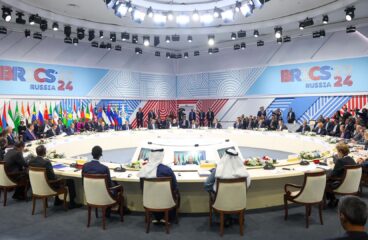 Reunión de la segunda sesión de la cumbre BRICS 2024 en Rusia, con representantes de varios países sentados en una mesa redonda, rodeados de banderas nacionales y bajo una intensa iluminación blanca. En el fondo, se aprecia el logo tricolor del evento. Mayoría mundial