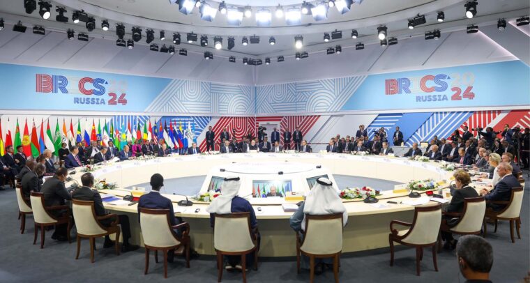 Reunión de la segunda sesión de la cumbre BRICS 2024 en Rusia, con representantes de varios países sentados en una mesa redonda, rodeados de banderas nacionales y bajo una intensa iluminación blanca. En el fondo, se aprecia el logo tricolor del evento. Mayoría mundial