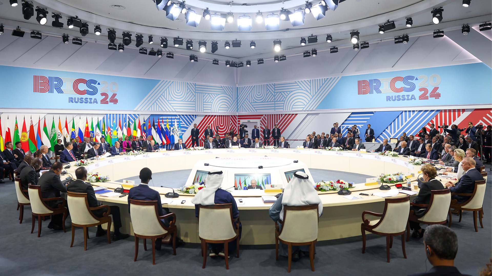 Reunión de la segunda sesión de la cumbre BRICS 2024 en Rusia, con representantes de varios países sentados en una mesa redonda, rodeados de banderas nacionales y bajo una intensa iluminación blanca. En el fondo, se aprecia el logo tricolor del evento. Mayoría mundial