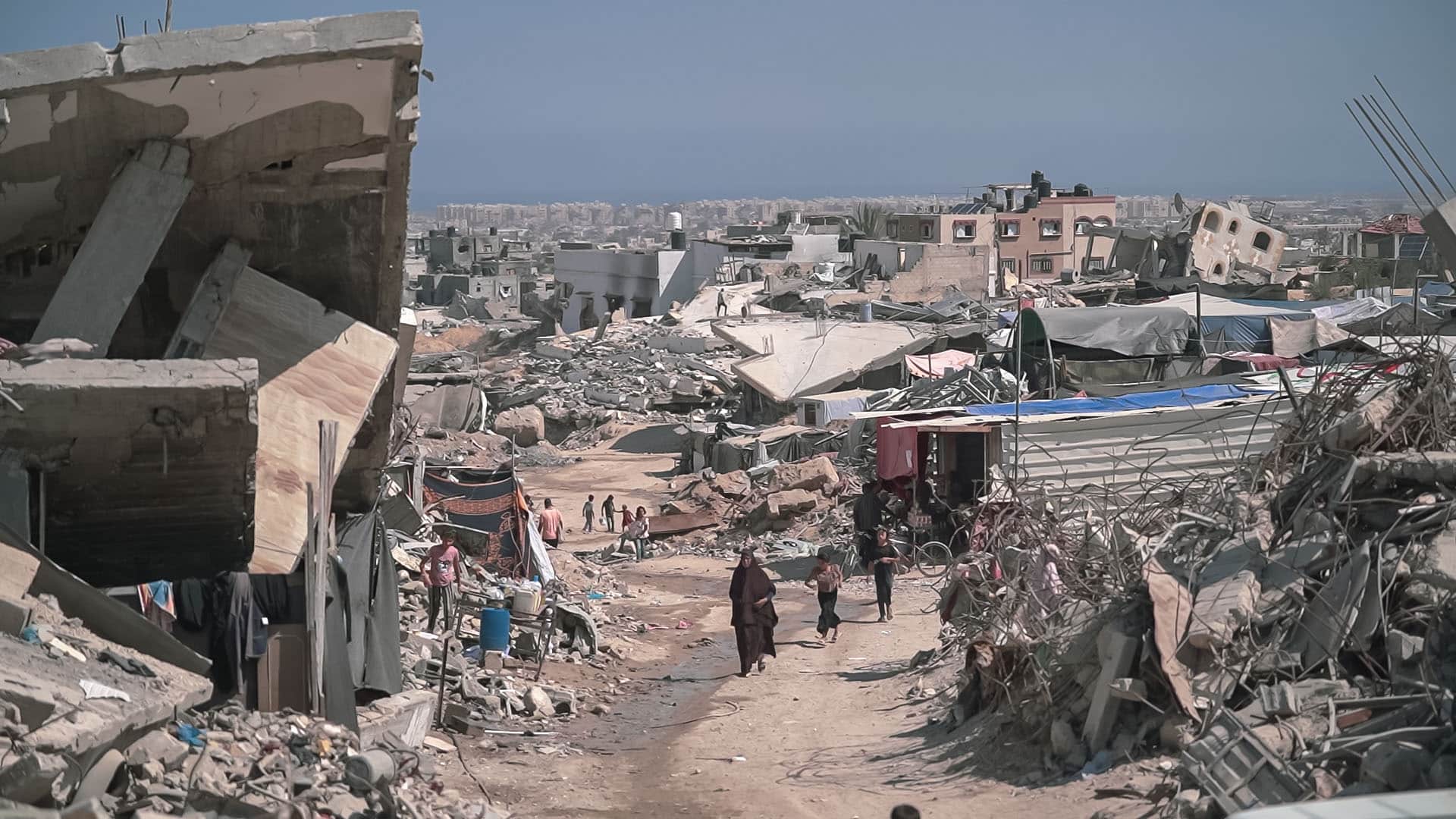 Zona urbana devastada en Gaza con escombros, edificios colapsados y personas caminando entre las ruinas. Gaza