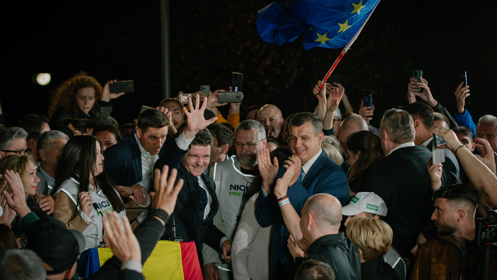 El candidato presidencial Nicușor Dan saluda a sus seguidores tras conocerse los primeros resultados de las encuestas a pie de urna durante la segunda vuelta de las elecciones presidenciales de Rumanía el 19 de mayo de 2025. Entre la multitud se ven una bandera de la Unión Europea y una de Rumanía. Varias personas capturan el momento con sus móviles. Rumanía