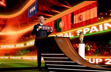 El presidente de la FIFA, Gianni Infantino, de pie sobre una tarima y vestido con traje oscuro y camisa blanca, sostiene una tarjeta negra con el texto “FIFA World Cup 2030” junto a los nombres y banderas de Marruecos, España y Portugal, anunciando oficialmente su designación como países anfitriones. A su derecha, el trofeo de la Copa Mundial se exhibe en un pedestal acristalado, destacado por una iluminación cenital. Al fondo, una pantalla LED proyecta los mismos elementos gráficos. Mundial 2030