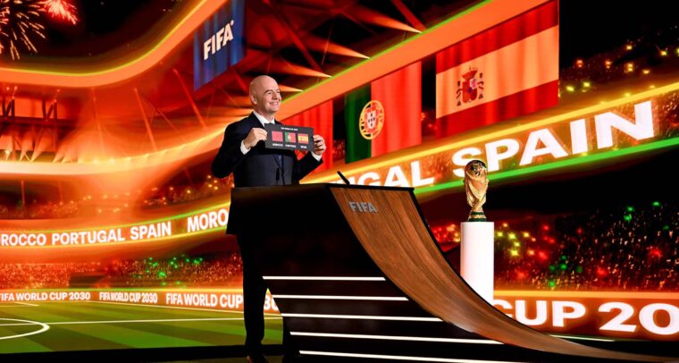 El presidente de la FIFA, Gianni Infantino, de pie sobre una tarima y vestido con traje oscuro y camisa blanca, sostiene una tarjeta negra con el texto “FIFA World Cup 2030” junto a los nombres y banderas de Marruecos, España y Portugal, anunciando oficialmente su designación como países anfitriones. A su derecha, el trofeo de la Copa Mundial se exhibe en un pedestal acristalado, destacado por una iluminación cenital. Al fondo, una pantalla LED proyecta los mismos elementos gráficos. Mundial 2030