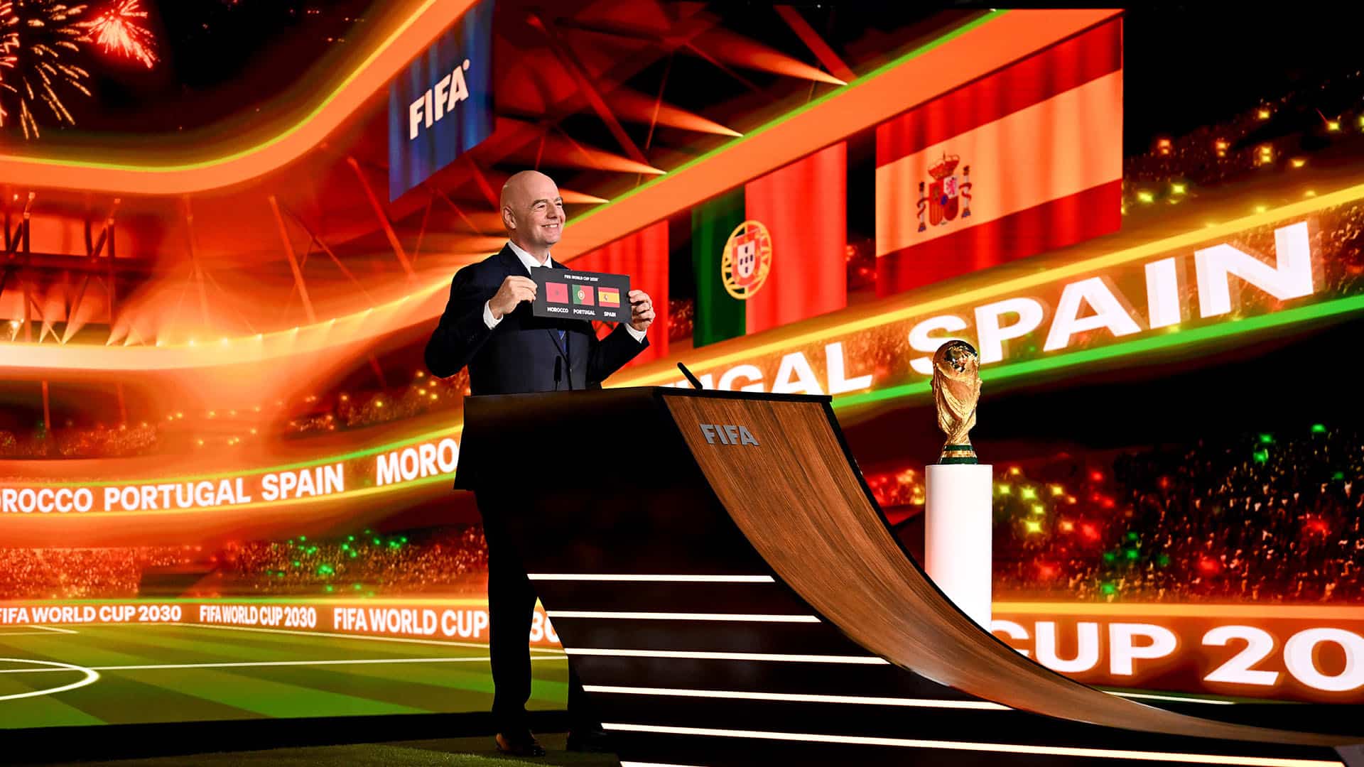 El presidente de la FIFA, Gianni Infantino, de pie sobre una tarima y vestido con traje oscuro y camisa blanca, sostiene una tarjeta negra con el texto “FIFA World Cup 2030” junto a los nombres y banderas de Marruecos, España y Portugal, anunciando oficialmente su designación como países anfitriones. A su derecha, el trofeo de la Copa Mundial se exhibe en un pedestal acristalado, destacado por una iluminación cenital. Al fondo, una pantalla LED proyecta los mismos elementos gráficos. Mundial 2030