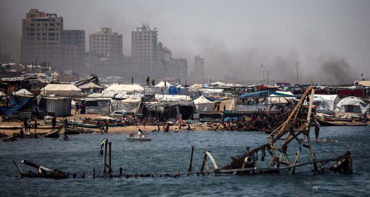 A line of tents near the port of Gaza, with several people gathered along the shoreline. In the background, buildings can be seen with smoke rising behind them. In the foreground, a partially submerged structure is visible in the water. Gaza
