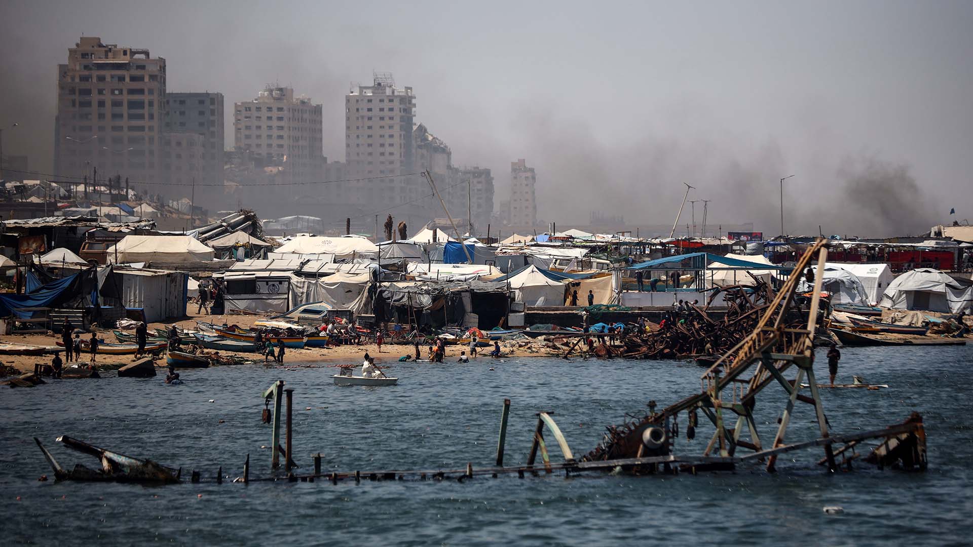 A line of tents near the port of Gaza, with several people gathered along the shoreline. In the background, buildings can be seen with smoke rising behind them. In the foreground, a partially submerged structure is visible in the water. Gaza
