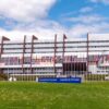 Vista frontal del Palacio de Europa en Estrasburgo, sede del Consejo de Europa, bajo cielo parcialmente nublado. El edificio, de hormigón y cristal, presenta una estructura simétrica con hileras de ventanales. Al frente, banderas de los Estados miembros se alinean sobre un amplio césped. A la derecha, se observa vegetación con varios árboles. Consejo de Europa