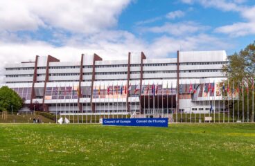 Vista frontal del Palacio de Europa en Estrasburgo, sede del Consejo de Europa, bajo cielo parcialmente nublado. El edificio, de hormigón y cristal, presenta una estructura simétrica con hileras de ventanales. Al frente, banderas de los Estados miembros se alinean sobre un amplio césped. A la derecha, se observa vegetación con varios árboles. Consejo de Europa