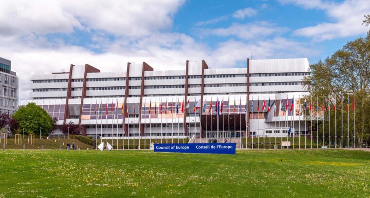 Vista frontal del Palacio de Europa en Estrasburgo, sede del Consejo de Europa, bajo cielo parcialmente nublado. El edificio, de hormigón y cristal, presenta una estructura simétrica con hileras de ventanales. Al frente, banderas de los Estados miembros se alinean sobre un amplio césped. A la derecha, se observa vegetación con varios árboles. Consejo de Europa