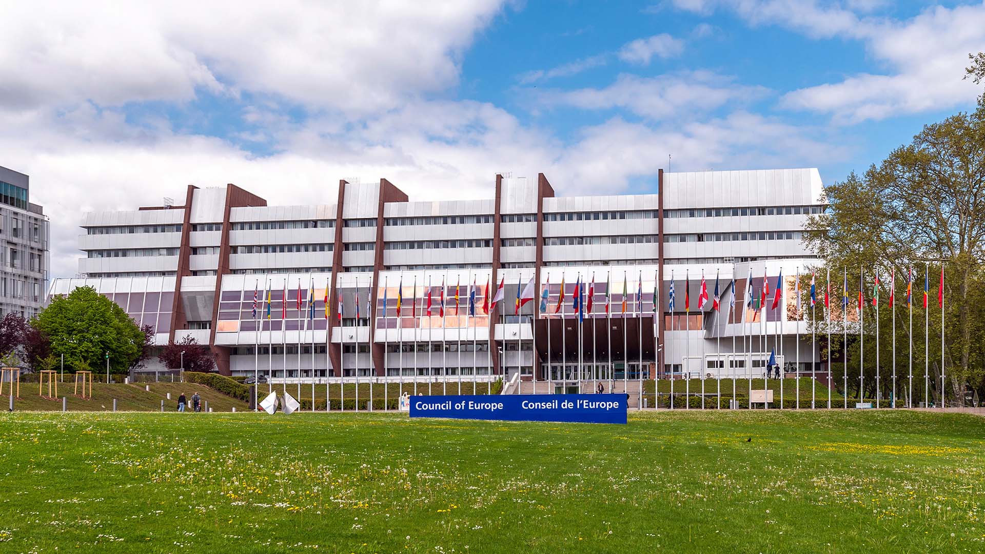 Vista frontal del Palacio de Europa en Estrasburgo, sede del Consejo de Europa, bajo cielo parcialmente nublado. El edificio, de hormigón y cristal, presenta una estructura simétrica con hileras de ventanales. Al frente, banderas de los Estados miembros se alinean sobre un amplio césped. A la derecha, se observa vegetación con varios árboles. Consejo de Europa
