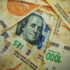 Detalle de un billete de 100 dólares estadounidenses, de tonos verdes y negros, parcialmente visible bajo varios billetes de 1.000 pesos argentinos en colores naranja, violeta y verde. Argentina