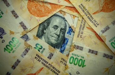 Detalle de un billete de 100 dólares estadounidenses, de tonos verdes y negros, parcialmente visible bajo varios billetes de 1.000 pesos argentinos en colores naranja, violeta y verde. Argentina