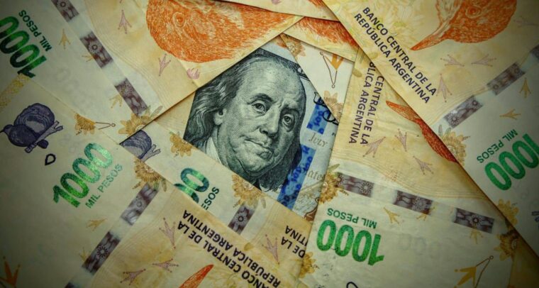 Detalle de un billete de 100 dólares estadounidenses, de tonos verdes y negros, parcialmente visible bajo varios billetes de 1.000 pesos argentinos en colores naranja, violeta y verde. Argentina