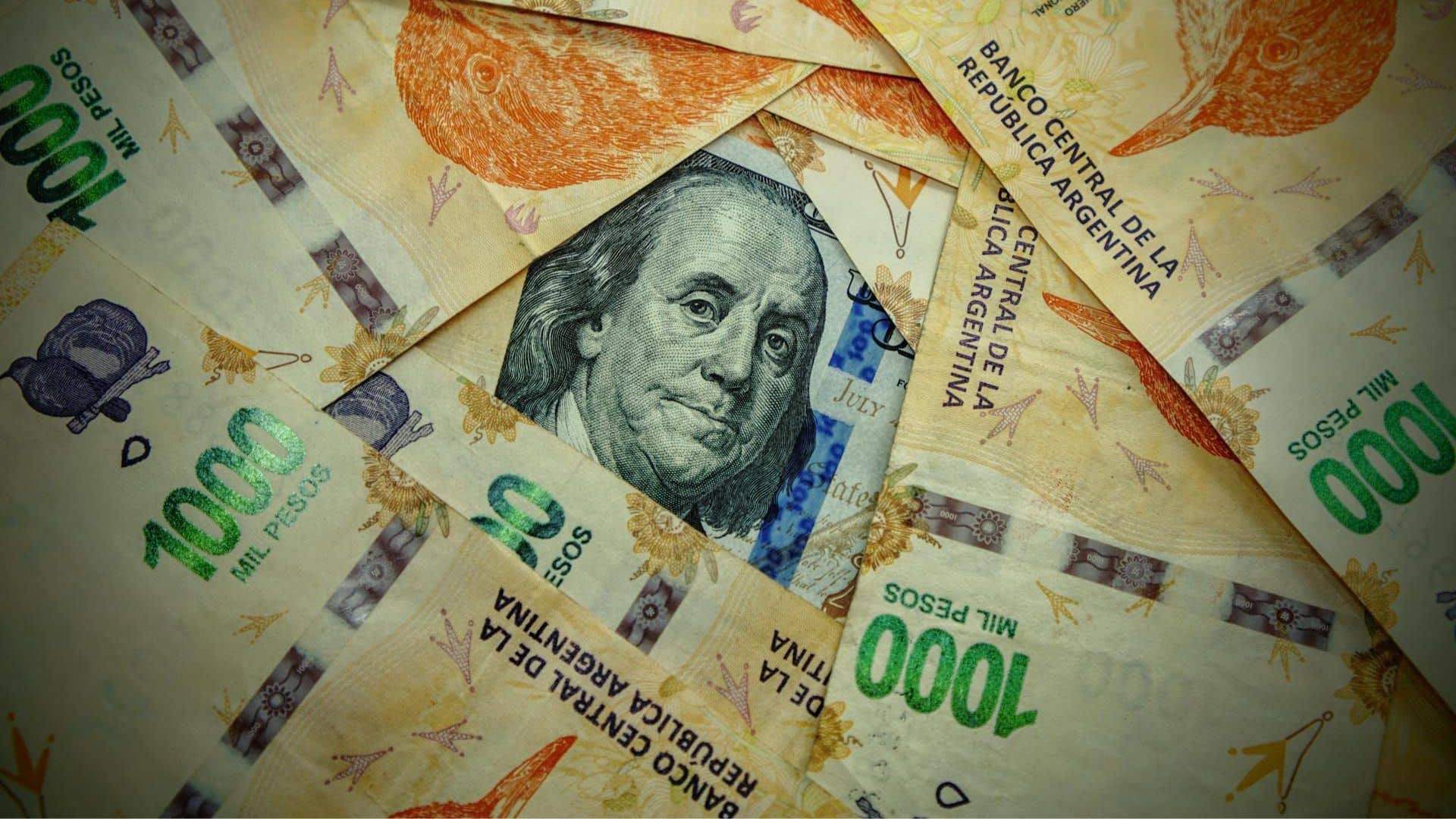 Detalle de un billete de 100 dólares estadounidenses, de tonos verdes y negros, parcialmente visible bajo varios billetes de 1.000 pesos argentinos en colores naranja, violeta y verde. Argentina