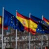 Banderas de España (rojo y amarillo con escudo) y de la Unión Europea (azul con estrellas doradas) ondeando juntas al viento frente a un edificio en un día soleado. Gabinetes