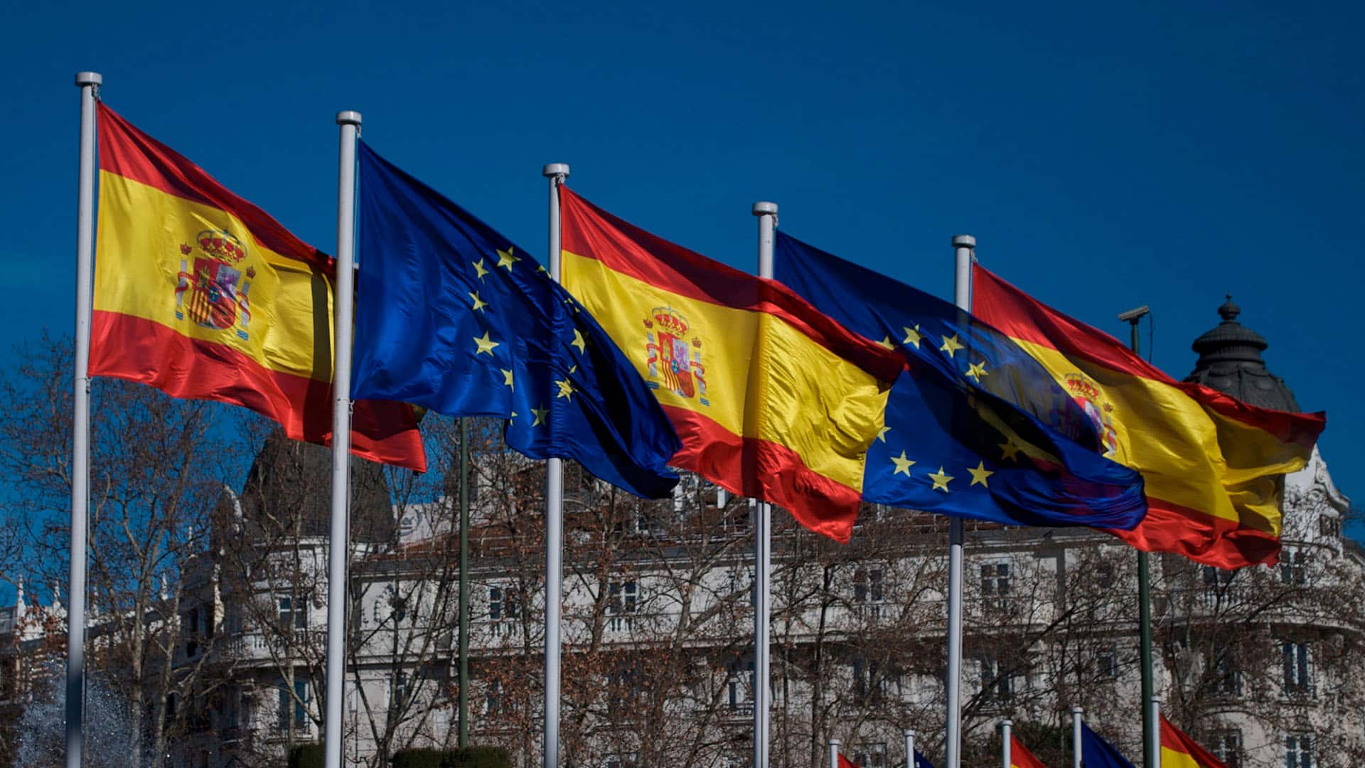 Banderas de España (rojo y amarillo con escudo) y de la Unión Europea (azul con estrellas doradas) ondeando juntas al viento frente a un edificio en un día soleado. Gabinetes