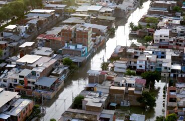 Vista aérea diurna de un vecindario urbano en Piura (Perú), afectado por inundaciones tras el paso de 'El Niño Costero' en 2017. Las calles están cubiertas de agua y algunas personas caminan por zonas anegadas. Las viviendas de varios pisos muestran daños visibles, mientras el nivel del agua permanece alto. América Latina