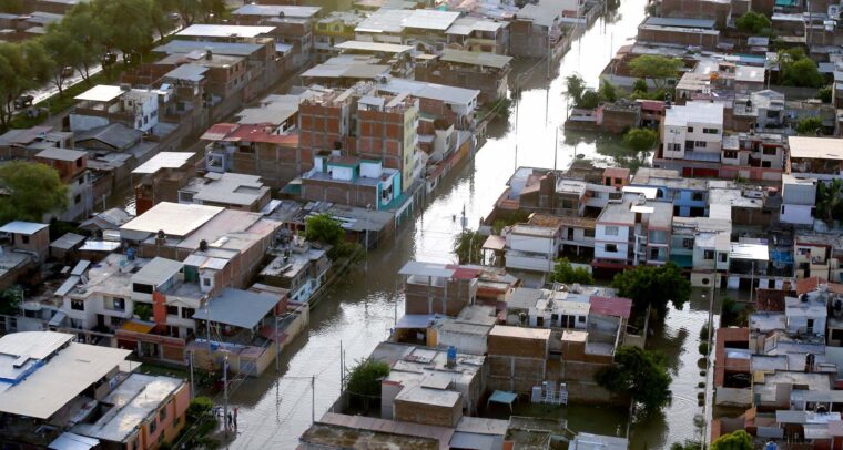 Vista aérea diurna de un vecindario urbano en Piura (Perú), afectado por inundaciones tras el paso de 'El Niño Costero' en 2017. Las calles están cubiertas de agua y algunas personas caminan por zonas anegadas. Las viviendas de varios pisos muestran daños visibles, mientras el nivel del agua permanece alto. América Latina