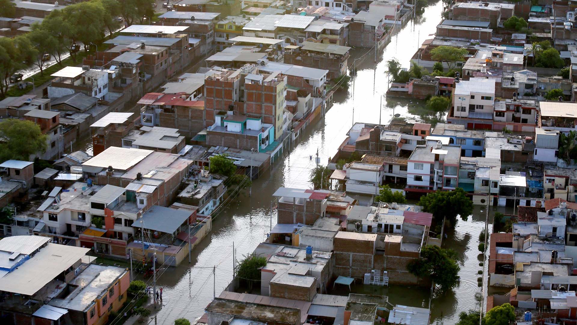 Vista aérea diurna de un vecindario urbano en Piura (Perú), afectado por inundaciones tras el paso de 'El Niño Costero' en 2017. Las calles están cubiertas de agua y algunas personas caminan por zonas anegadas. Las viviendas de varios pisos muestran daños visibles, mientras el nivel del agua permanece alto. América Latina