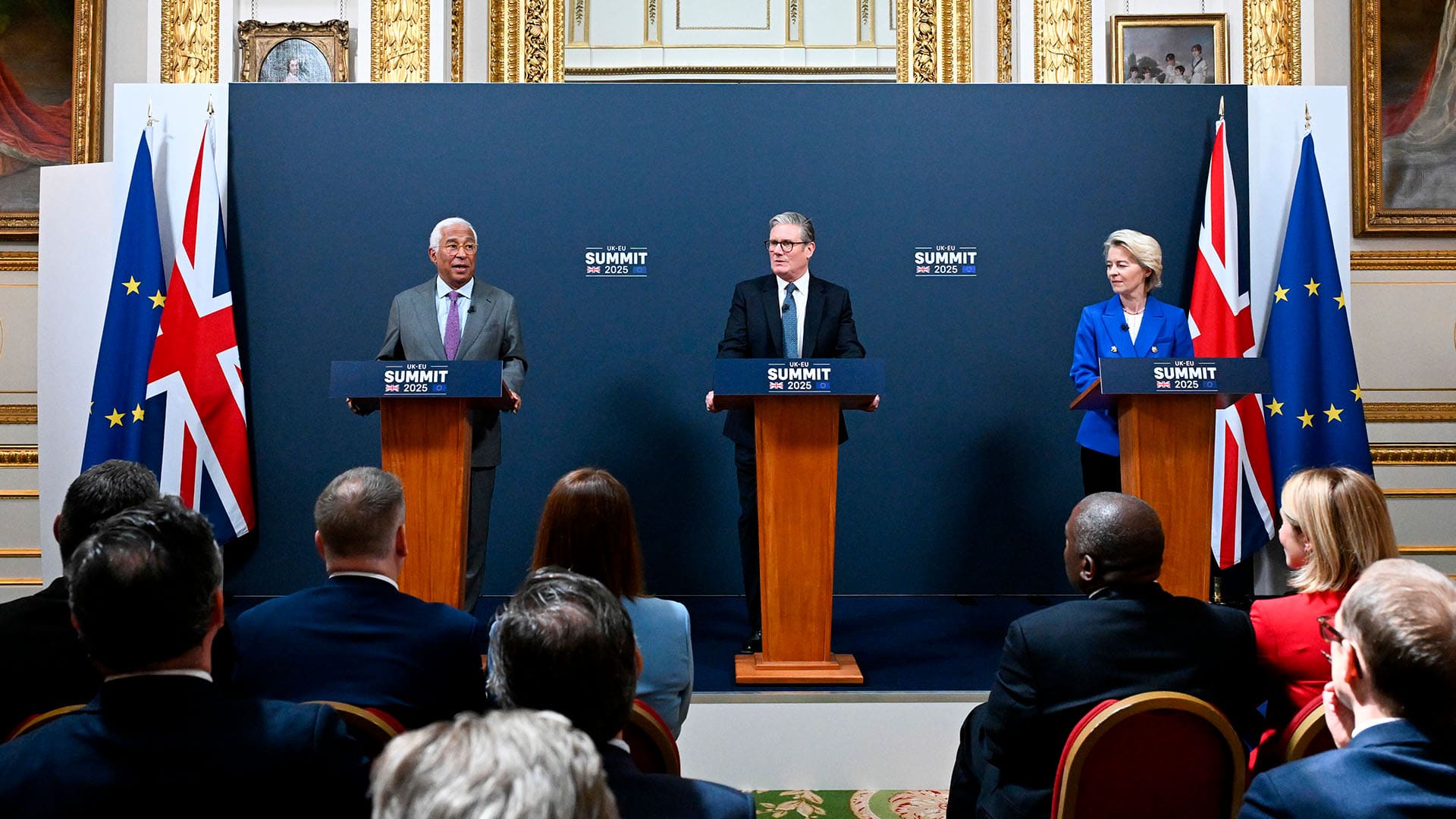 De izquierda a derecha, António Costa (con traje gris, camisa azul clara y corbata lila), presidente del Consejo Europeo; Keir Starmer (con traje negro, camisa blanca y corbata azul), primer ministro del Reino Unido; y Ursula von der Leyen (con chaqueta azul, camisa blanca y pantalones negros), presidenta de la Comisión Europea, de pie detrás de atriles con el rótulo "UK-EU Summit 2025", durante una rueda de prensa conjunta celebrada el 19 de mayo de 2025 en una sala con ornamentación dorada, pinturas clásicas y banderas de la UE y del Reino Unido al fondo. Frente a ellos, un grupo de personas los escucha con atención. Reino Unido