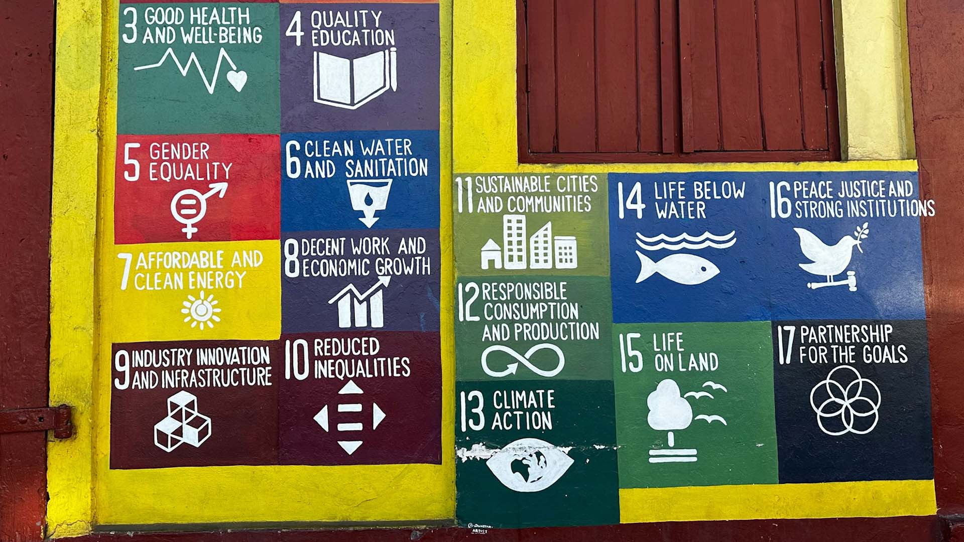 Mural pintado en una pared que representa los 17 Objetivos de Desarrollo Sostenible (ODS) de las Naciones Unidas, cada uno con un ícono y color distintivo. A la izquierda del mural hay una puerta roja con un cartel que dice "NO VENDING". A la derecha, sobre el mural, se encuentra una ventana de madera color terracota, enmarcada por una franja amarilla. Desarrollo