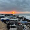 Campamento improvisado de personas desplazadas en la costa de la Franja de Gaza al atardecer, con refugios y tiendas hechas de lonas y materiales diversos, agrupados bajo un cielo nublado. Al fondo se ve el mar y el sol parcialmente oculto tras las nubes grises. Gaza