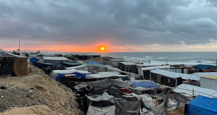 Improvised camp for displaced people on the coast of the Gaza Strip at sunset, with tents and shelters made of tarpaulins and assorted materials, clustered closely under a cloudy sky. In the background, the sea and the sun are partially hidden behind grey clouds. Gaza