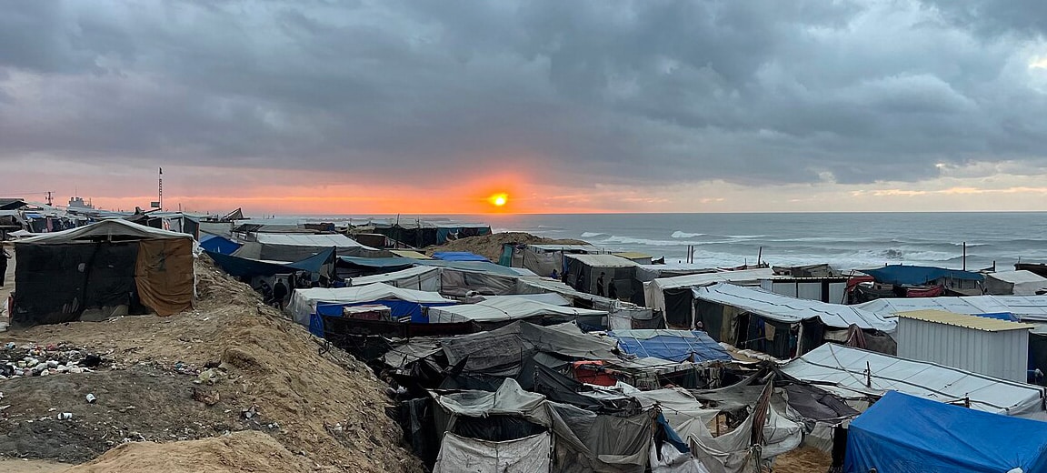 Campamento improvisado de personas desplazadas en la costa de la Franja de Gaza al atardecer, con refugios y tiendas hechas de lonas y materiales diversos, agrupados bajo un cielo nublado. Al fondo se ve el mar y el sol parcialmente oculto tras las nubes grises. Gaza