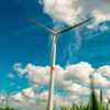 Aerogenerador en primer plano en un campo verde en Villers-le-Bouillet, cerca de Lieja (Bélgica), bajo un cielo azul con nubes, con más turbinas eólicas visibles en el horizonte. Energética