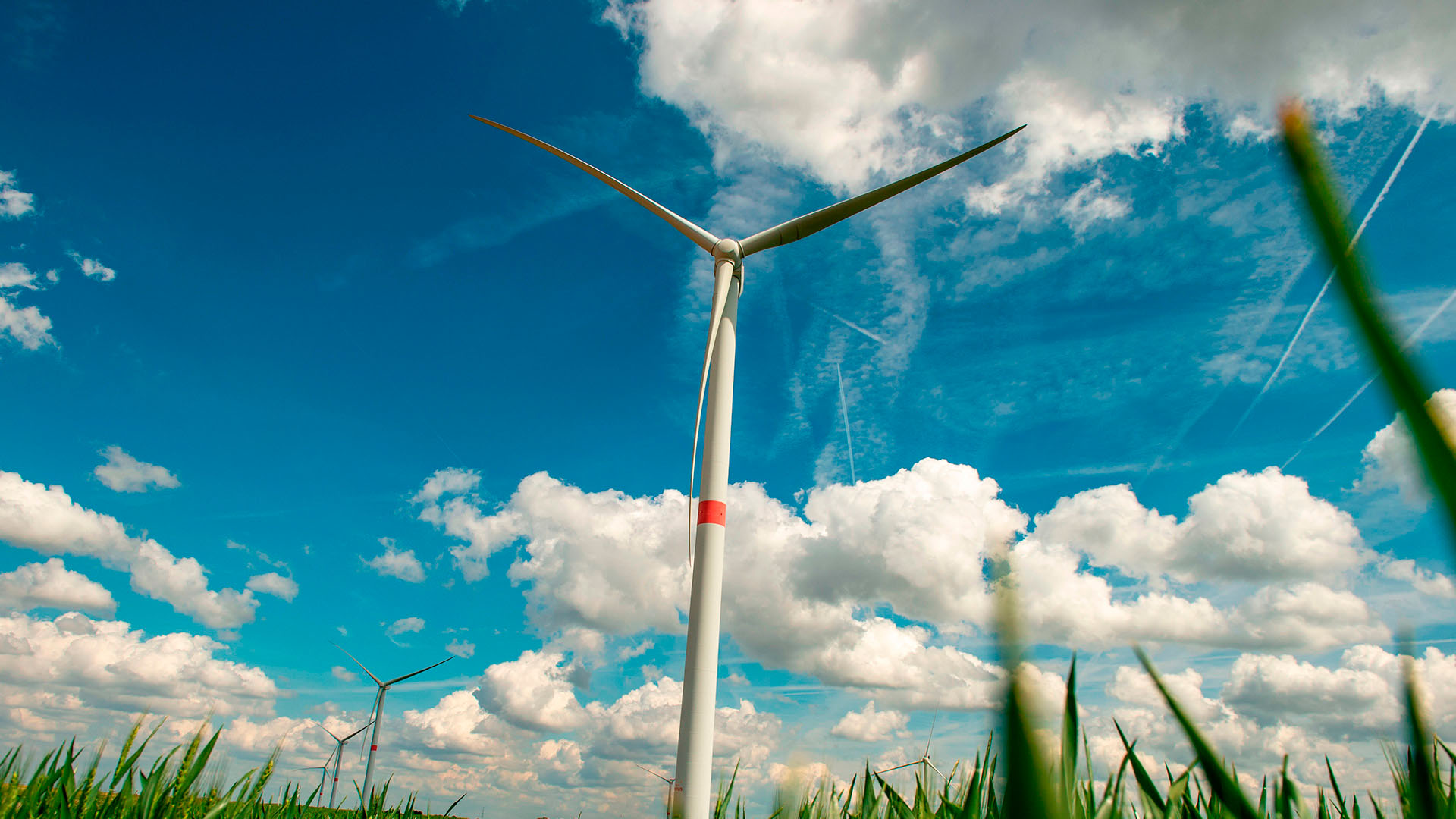 Aerogenerador en primer plano en un campo verde en Villers-le-Bouillet, cerca de Lieja (Bélgica), bajo un cielo azul con nubes, con más turbinas eólicas visibles en el horizonte. Energética