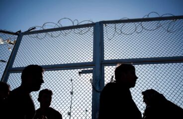 Siluetas de varias personas migrantes que esperan frente a una valla con alambre de espino en un centro de registro de un campo de refugiados en Lesbos (Grecia), en un día soleado. Migración