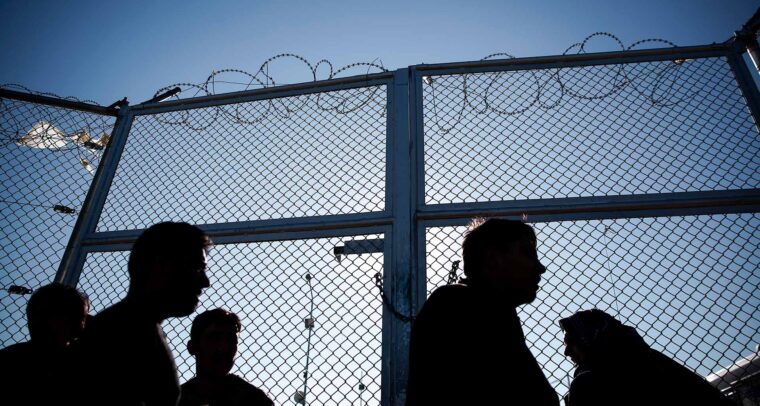 Siluetas de varias personas migrantes que esperan frente a una valla con alambre de espino en un centro de registro de un campo de refugiados en Lesbos (Grecia), en un día soleado. Migración