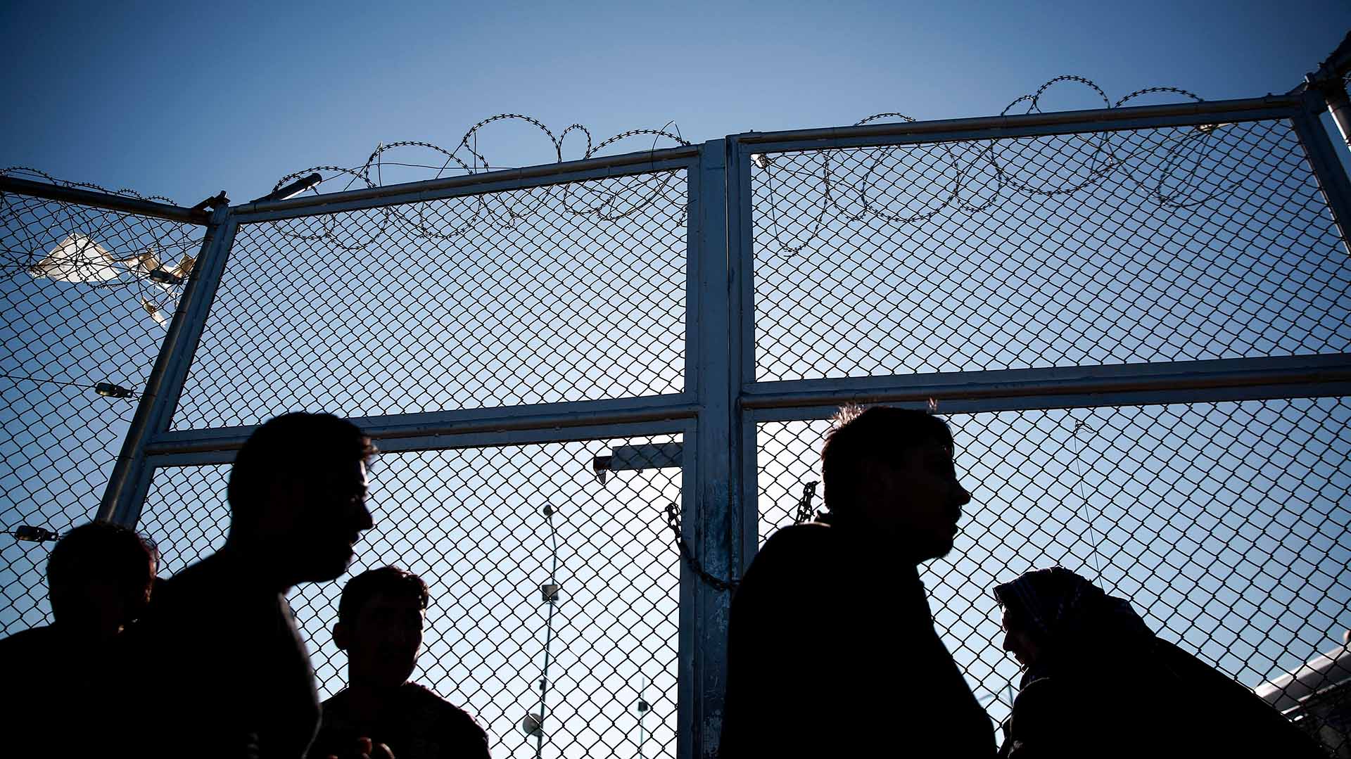 Siluetas de varias personas migrantes que esperan frente a una valla con alambre de espino en un centro de registro de un campo de refugiados en Lesbos (Grecia), en un día soleado. Migración