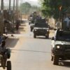 Convoy de vehículos militares 4x4 pertenecientes a la unidad de protección EUTM-Mali, patrullando durante el día entre las localidades de Bamako y Koulikoro (Malí). En primer plano, una persona se desplaza en un carro tirado por un burro, mientras varias personas caminan por la calle en el lado opuesto a los vehículos. A la izquierda, se aprecian edificaciones bajas de adobe. Sahel