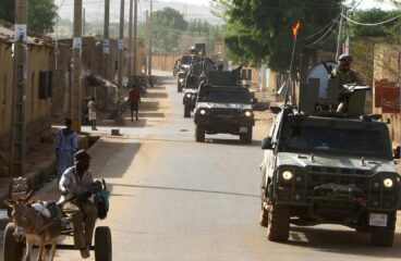 Convoy de vehículos militares 4x4 pertenecientes a la unidad de protección EUTM-Mali, patrullando durante el día entre las localidades de Bamako y Koulikoro (Malí). En primer plano, una persona se desplaza en un carro tirado por un burro, mientras varias personas caminan por la calle en el lado opuesto a los vehículos. A la izquierda, se aprecian edificaciones bajas de adobe. Sahel