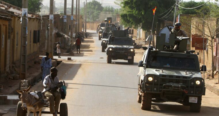 Convoy de vehículos militares 4x4 pertenecientes a la unidad de protección EUTM-Mali, patrullando durante el día entre las localidades de Bamako y Koulikoro (Malí). En primer plano, una persona se desplaza en un carro tirado por un burro, mientras varias personas caminan por la calle en el lado opuesto a los vehículos. A la izquierda, se aprecian edificaciones bajas de adobe. Sahel