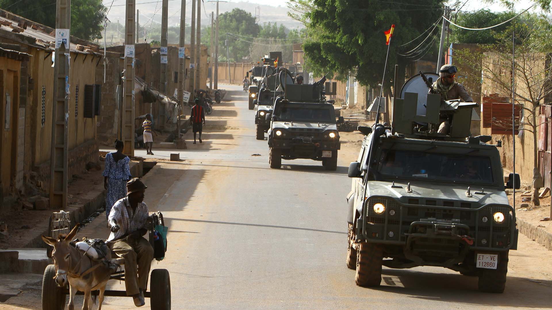 Convoy de vehículos militares 4x4 pertenecientes a la unidad de protección EUTM-Mali, patrullando durante el día entre las localidades de Bamako y Koulikoro (Malí). En primer plano, una persona se desplaza en un carro tirado por un burro, mientras varias personas caminan por la calle en el lado opuesto a los vehículos. A la izquierda, se aprecian edificaciones bajas de adobe. Sahel