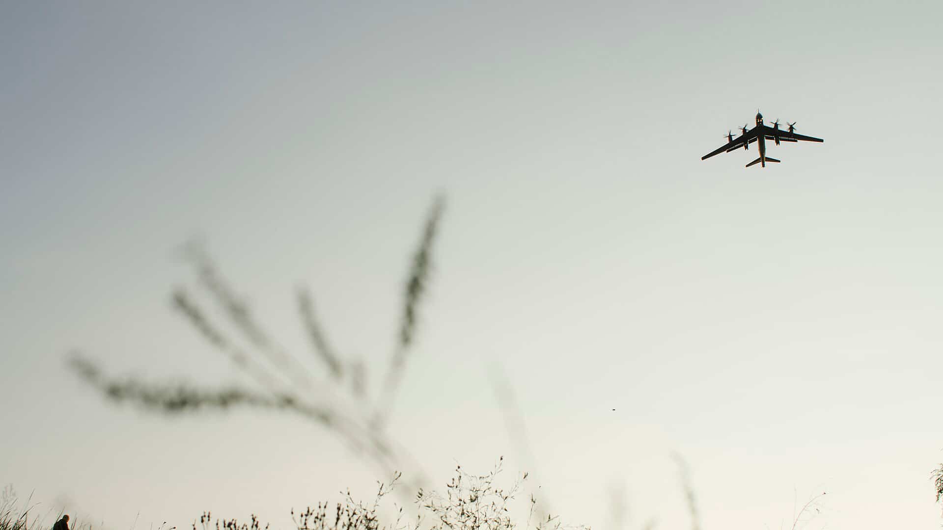 Silueta de un avión militar Tu-95 regresando a su base, volando a baja altura sobre un campo con vegetación al atardecer. A la izquierda, se distingue la silueta de una persona con una mochila. Ucrania