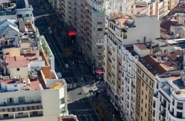 Vista aérea diurna de la Gran Vía en Madrid (España), en un día soleado. A ambos lados de la avenida destacan edificios de diferentes colores y estilos arquitectónicos. En el centro, la calzada y el área peatonal muestran vehículos y peatones desplazándose en diversas direcciones. España
