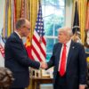 President Donald Trump greets German Chancellor Friedrich Merz with a handshake in the Oval Office. Both leaders are dressed in dark suits and stand in front of a display of American flags and military banners. A bust of Abraham Lincoln and framed photographs are visible in the background, along with large windows providing natural light. Germany