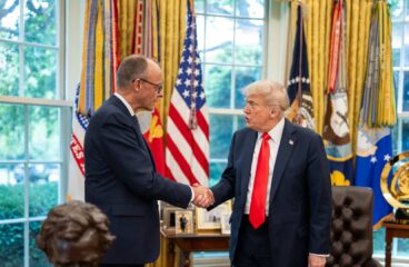 President Donald Trump greets German Chancellor Friedrich Merz with a handshake in the Oval Office. Both leaders are dressed in dark suits and stand in front of a display of American flags and military banners. A bust of Abraham Lincoln and framed photographs are visible in the background, along with large windows providing natural light. Germany