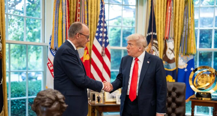 President Donald Trump greets German Chancellor Friedrich Merz with a handshake in the Oval Office. Both leaders are dressed in dark suits and stand in front of a display of American flags and military banners. A bust of Abraham Lincoln and framed photographs are visible in the background, along with large windows providing natural light. Germany