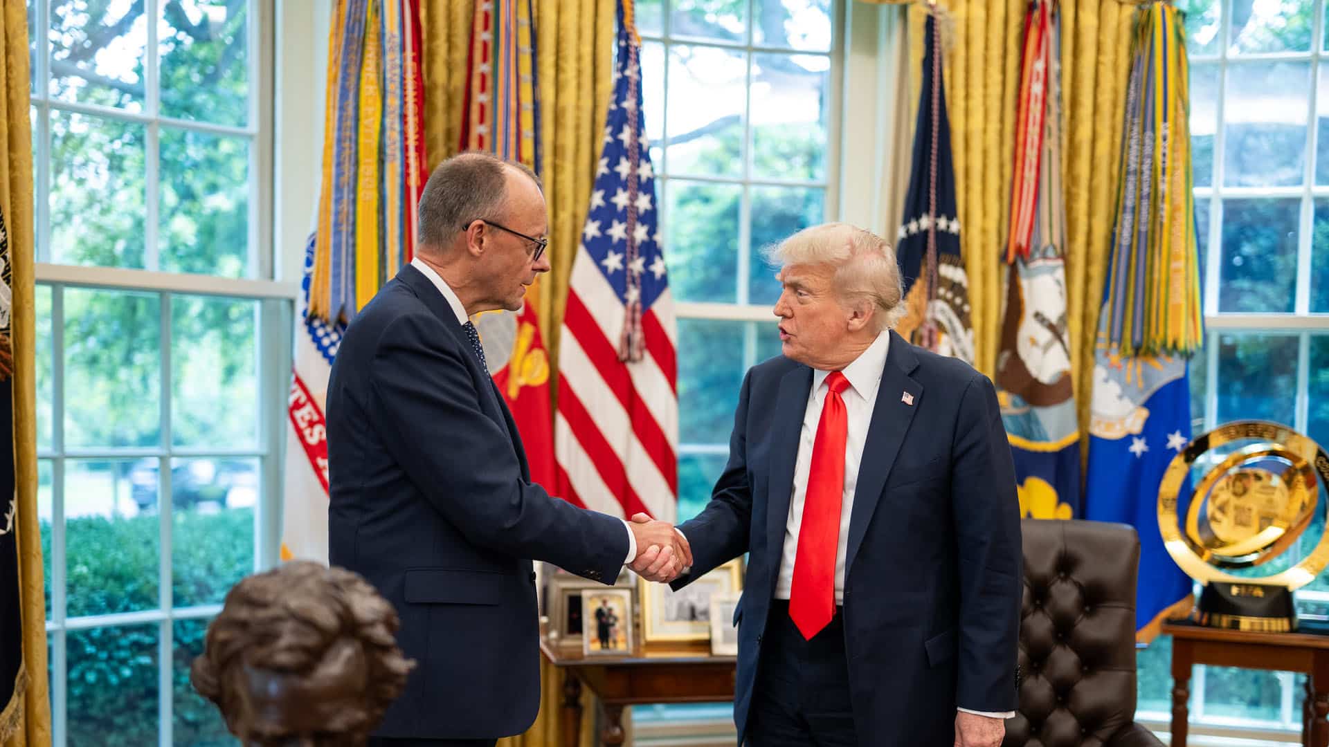 President Donald Trump greets German Chancellor Friedrich Merz with a handshake in the Oval Office. Both leaders are dressed in dark suits and stand in front of a display of American flags and military banners. A bust of Abraham Lincoln and framed photographs are visible in the background, along with large windows providing natural light. Germany