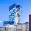 Edificio del Banco Central Europeo al atardecer, donde resalta la torre de cristal angular iluminada. En el lateral se observa una fachada de ladrillo. En el centro, la entrada de aluminio y cristal. Al frente, se observan varias farolas, así como la entrada al edificio. Trump