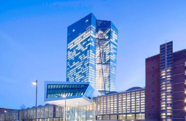 Edificio del Banco Central Europeo al atardecer, donde resalta la torre de cristal angular iluminada. En el lateral se observa una fachada de ladrillo. En el centro, la entrada de aluminio y cristal. Al frente, se observan varias farolas, así como la entrada al edificio. Trump