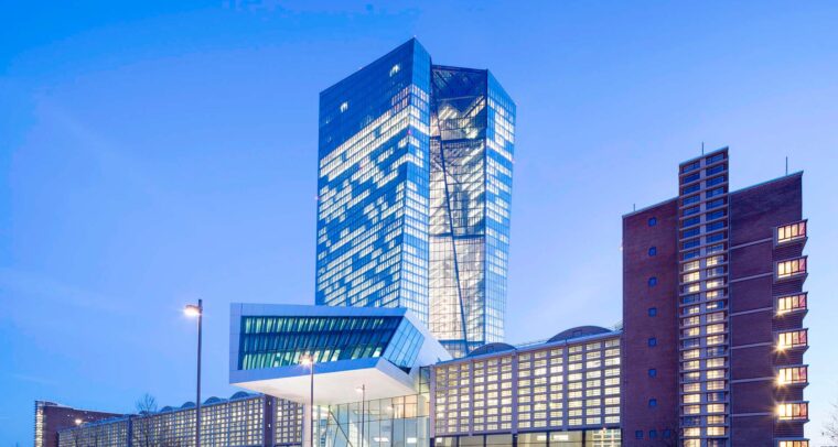 Edificio del Banco Central Europeo al atardecer, donde resalta la torre de cristal angular iluminada. En el lateral se observa una fachada de ladrillo. En el centro, la entrada de aluminio y cristal. Al frente, se observan varias farolas, así como la entrada al edificio. Trump