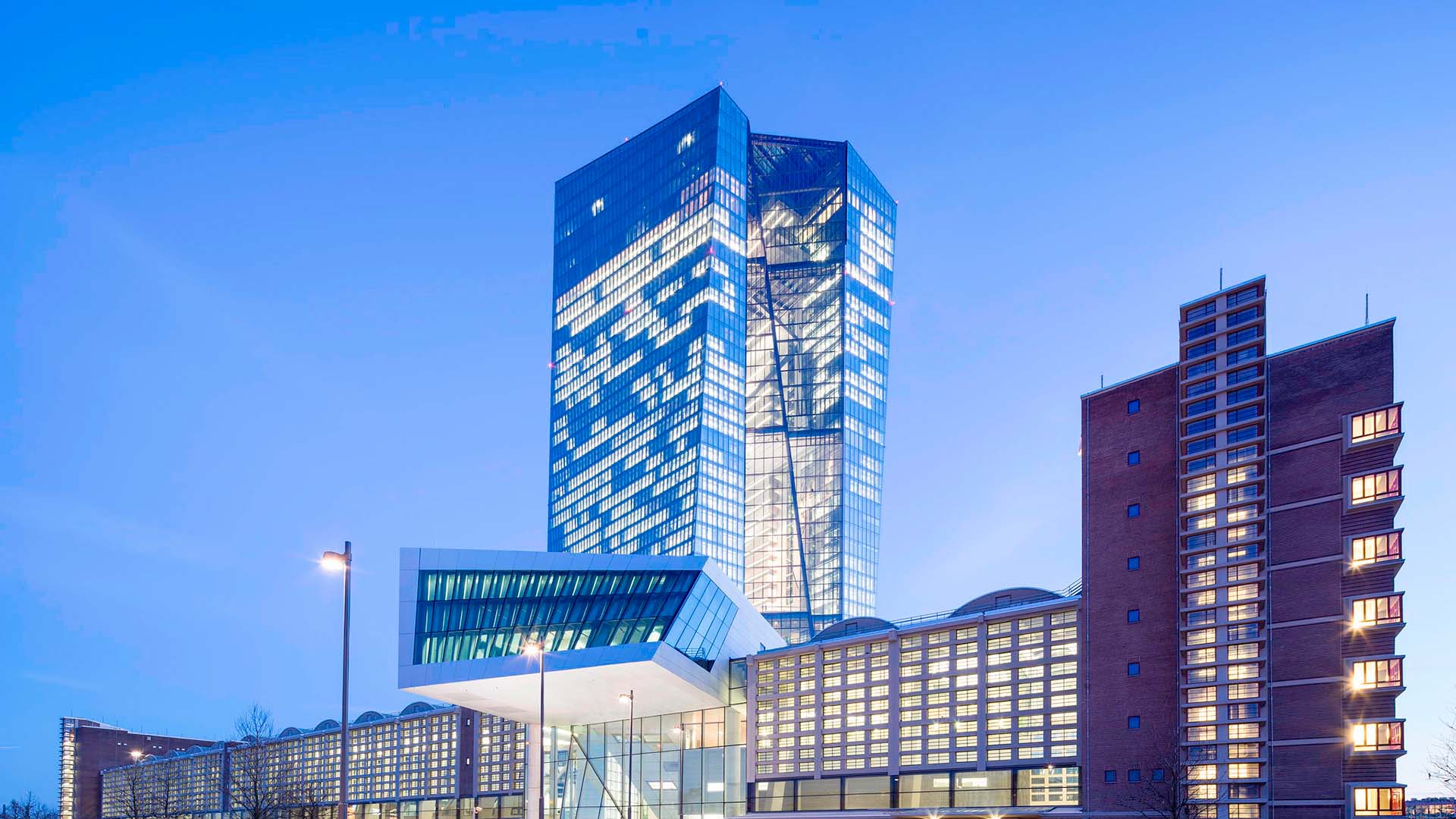 Edificio del Banco Central Europeo al atardecer, donde resalta la torre de cristal angular iluminada. En el lateral se observa una fachada de ladrillo. En el centro, la entrada de aluminio y cristal. Al frente, se observan varias farolas, así como la entrada al edificio. Trump
